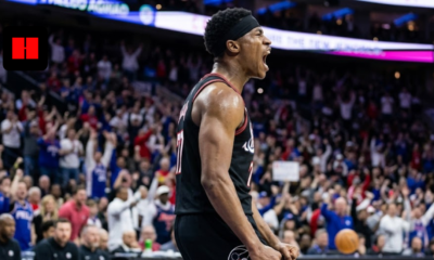 A professional basketball player in a black Philadelphia 76ers jersey and headband, captured from a side angle while shouting in celebration during a high-energy game with a blurred crowd in the background.
