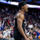 A professional basketball player in a black Philadelphia 76ers jersey and headband, captured from a side angle while shouting in celebration during a high-energy game with a blurred crowd in the background.