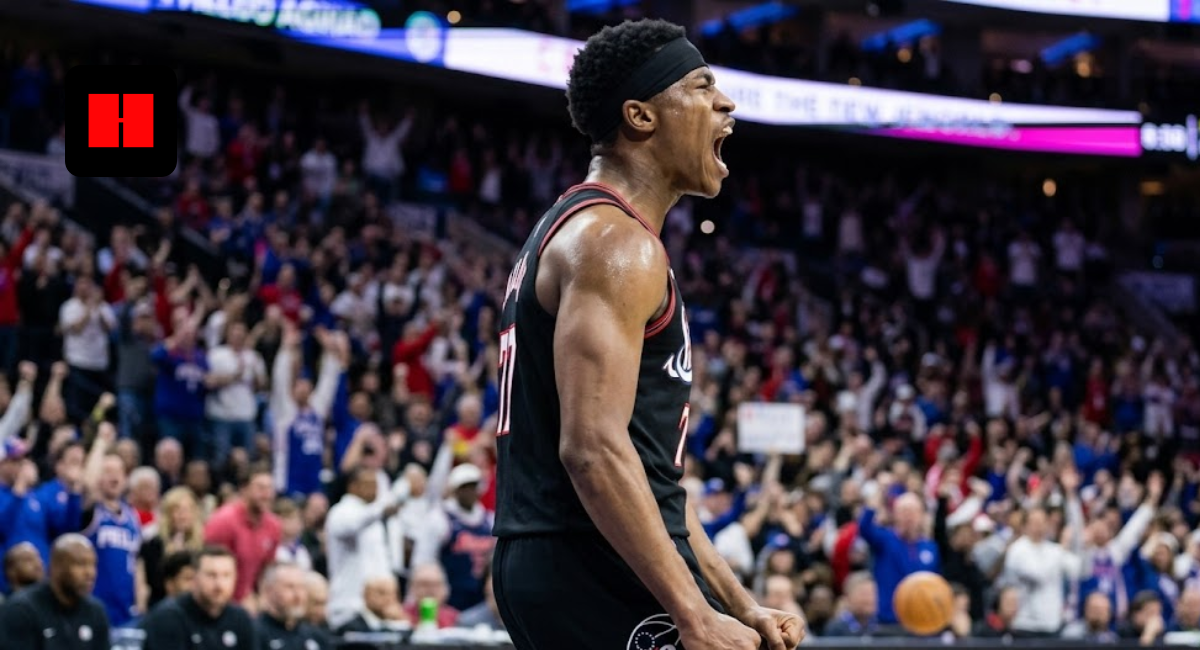 A professional basketball player in a black Philadelphia 76ers jersey and headband, captured from a side angle while shouting in celebration during a high-energy game with a blurred crowd in the background.