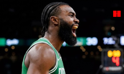 Jaylen Brown of the Boston Celtics shouting in celebration during a game, wearing a green jersey with white braided hair, side profile view in a dark arena.