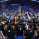 Wide-angle fisheye shot of the Oklahoma City Thunder players celebrating on a podium, holding the gold Larry O'Brien NBA Championship trophy amidst blue and gold confetti.