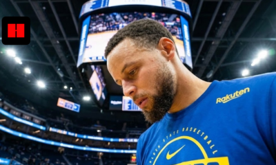 Stephen Curry looking down in a blue Golden State Warriors long-sleeve warmup shirt on the court at Chase Center.