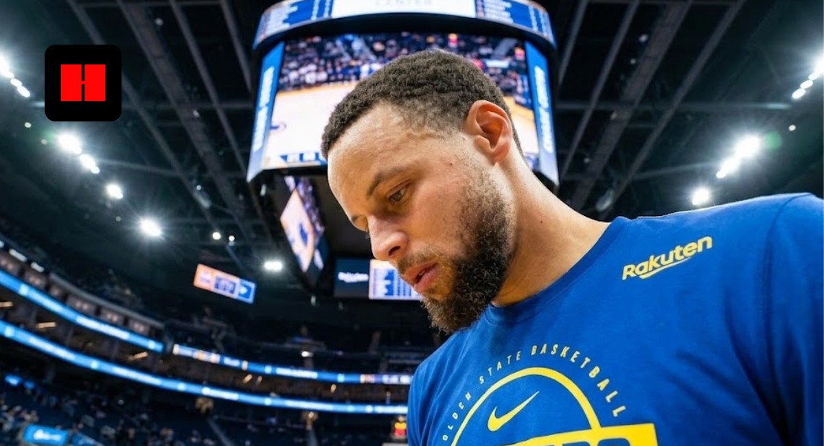 Stephen Curry looking down in a blue Golden State Warriors long-sleeve warmup shirt on the court at Chase Center.