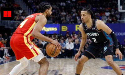 An Atlanta Hawks basketball player in a red and yellow jersey dribbles the ball while being closely guarded by a Detroit Pistons player in a black and teal jersey during an NBA game.