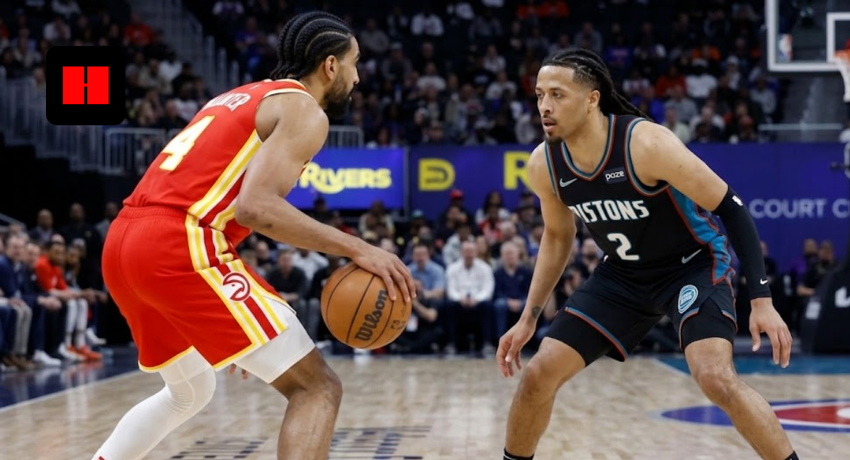 An Atlanta Hawks basketball player in a red and yellow jersey dribbles the ball while being closely guarded by a Detroit Pistons player in a black and teal jersey during an NBA game.