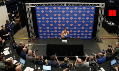 Wide high-angle shot of Jalen Brunson at a Knicks press conference surrounded by media.