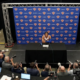 Wide high-angle shot of Jalen Brunson at a Knicks press conference surrounded by media.