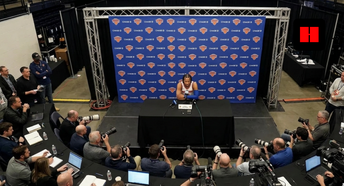 Wide high-angle shot of Jalen Brunson at a Knicks press conference surrounded by media.