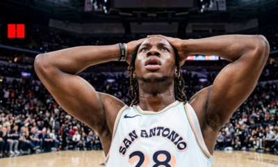A low-angle shot of San Antonio Spurs player Charles Bassey (number 28) with his hands on his head in a crowded basketball arena.