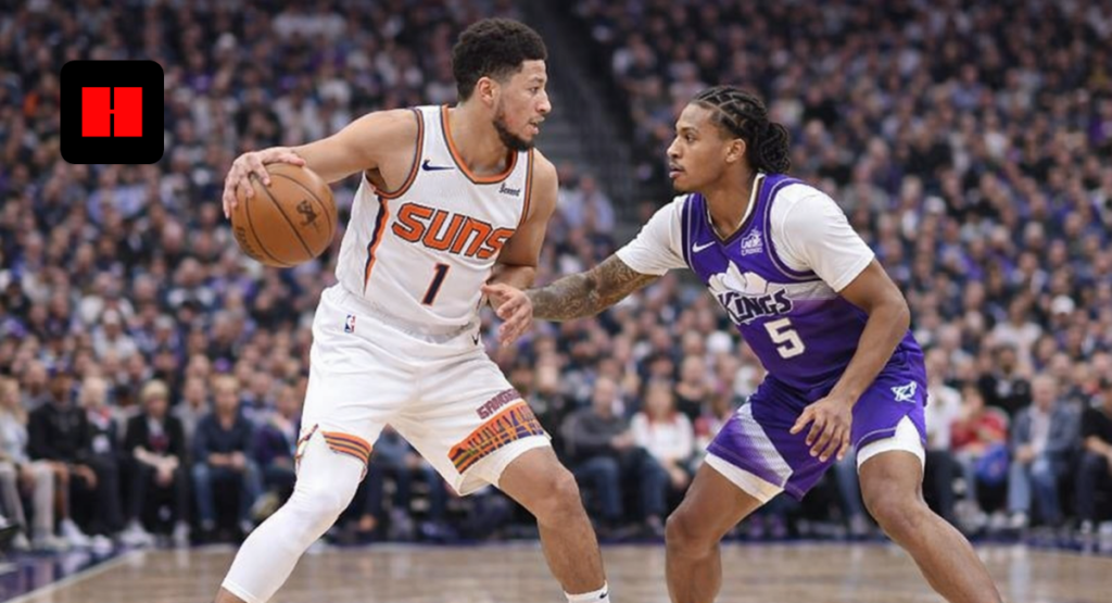 Devin Booker dribbling the basketball against De’Aaron Fox during an NBA game on the court with crowd in background.