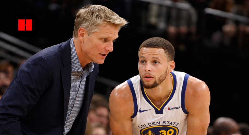 Golden State Warriors coach talking to player during NBA game strategy discussion captured from a side angle with arena crowd in background.