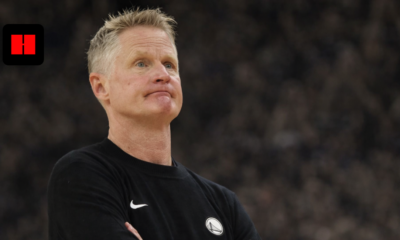 Golden State Warriors head coach standing with arms crossed, captured from a side camera angle during an NBA game with blurred arena crowd background