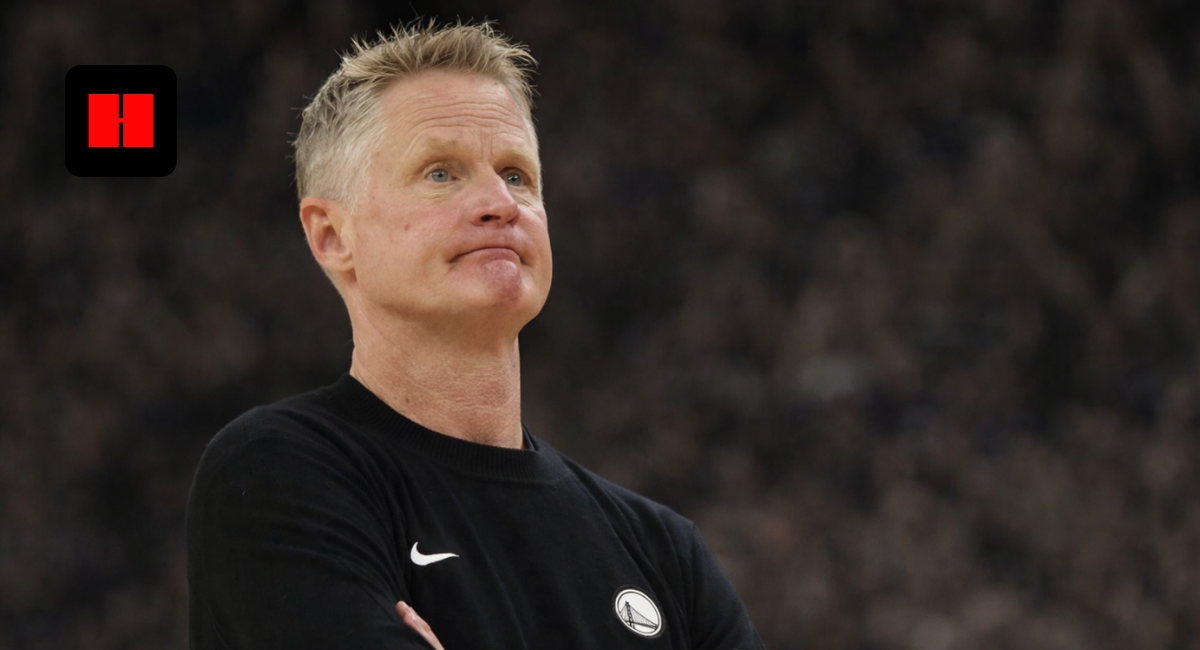 Golden State Warriors head coach standing with arms crossed, captured from a side camera angle during an NBA game with blurred arena crowd background
