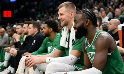 Boston Celtics stars Jaylen Brown and Kristaps Porzingis smiling and laughing while sitting on the team bench during an NBA game.