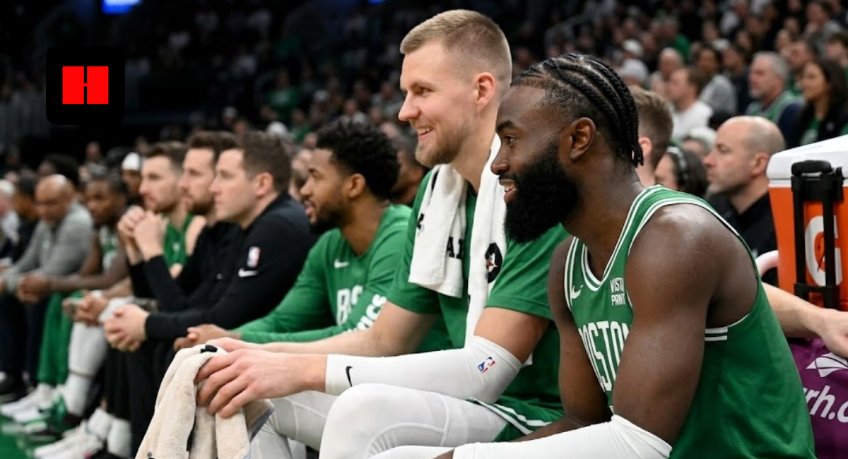 Boston Celtics stars Jaylen Brown and Kristaps Porzingis smiling and laughing while sitting on the team bench during an NBA game.