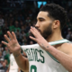 Jayson Tatum of the Boston Celtics holding his white jersey out in celebration during an NBA game, captured from a side angle with a blurred crowd in the background.
