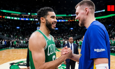 Two tall men, Jayson Tatum and Kristaps Porzingis, in green and blue basketball jerseys respectively, smiling as they shake hands on a crowded basketball court.