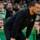 Boston Celtics head coach Joe Mazzulla leaning forward intensely on the sidelines during an NBA game, wearing a black crewneck sweatshirt with a small cross detail.