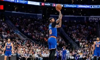 NBA player Jordan Clarkson in a blue New York Knicks jersey performing a jump shot during a basketball game in a crowded arena.
