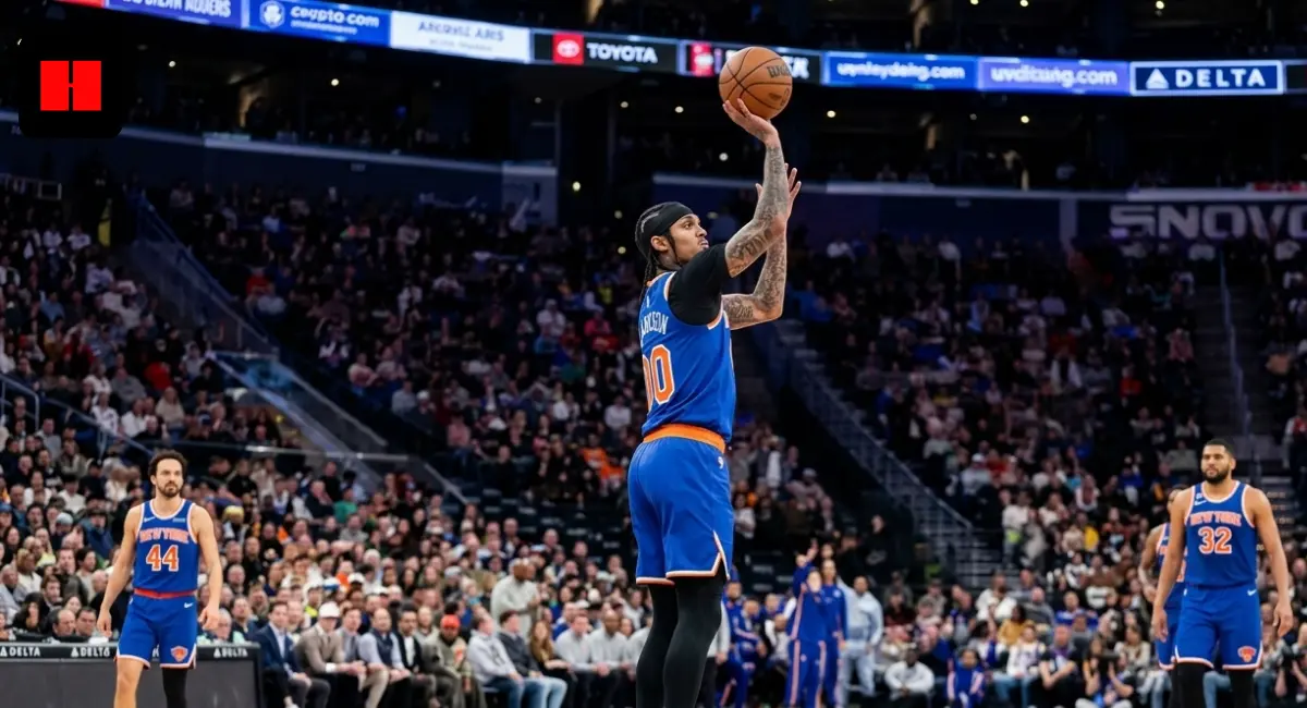 NBA player Jordan Clarkson in a blue New York Knicks jersey performing a jump shot during a basketball game in a crowded arena.