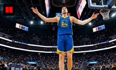 Luka Dončić in a Golden State Warriors uniform stands under the basket with arms raised during a home game, crowd and scoreboard visible in the background.