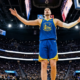 Luka Dončić in a Golden State Warriors uniform stands under the basket with arms raised during a home game, crowd and scoreboard visible in the background.