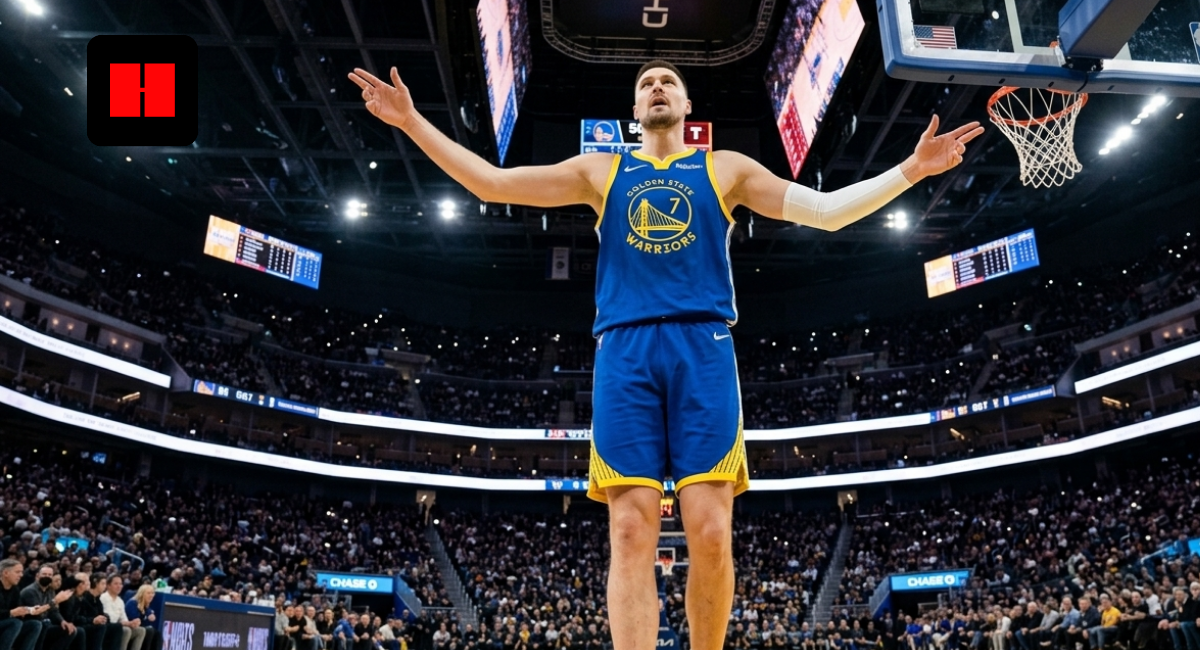 Luka Dončić in a Golden State Warriors uniform stands under the basket with arms raised during a home game, crowd and scoreboard visible in the background.