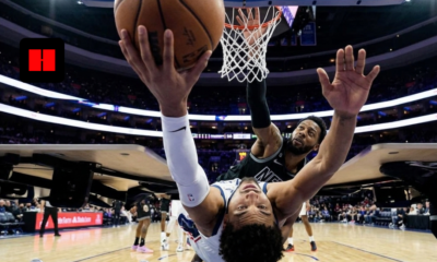 NBA player attempting a reverse layup under the basket while a defender contests the shot during a live game inside a packed arena.