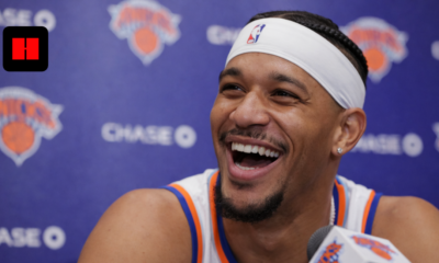 New York Knicks player smiling during an NBA press conference with team backdrop and microphone