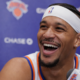 New York Knicks player smiling during an NBA press conference with team backdrop and microphone