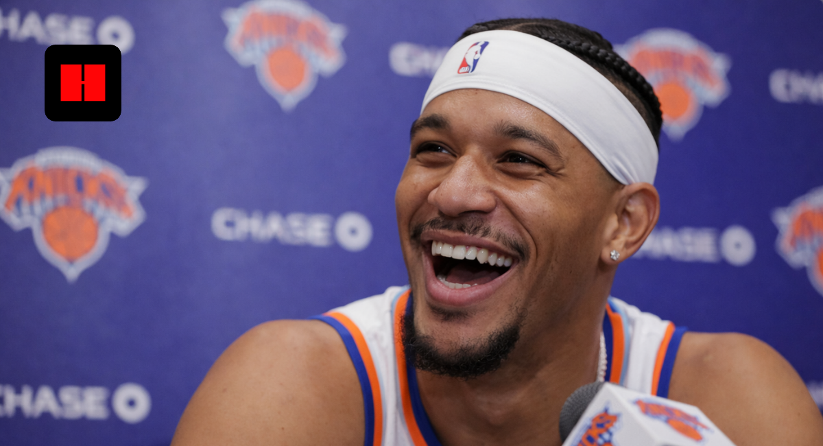 New York Knicks player smiling during an NBA press conference with team backdrop and microphone