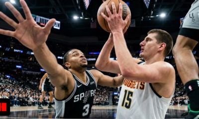 Low-angle floor-cam photo from below the rim showing Denver Nuggets center Nikola Jokic lifting a shot over San Antonio Spurs defender Jordan Hall, highlighting the close proximity and massive reaching arm of the defender.