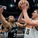 Low-angle floor-cam photo from below the rim showing Denver Nuggets center Nikola Jokic lifting a shot over San Antonio Spurs defender Jordan Hall, highlighting the close proximity and massive reaching arm of the defender.