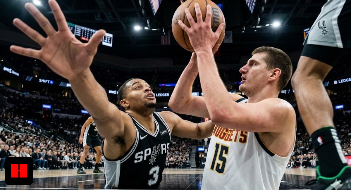 Low-angle floor-cam photo from below the rim showing Denver Nuggets center Nikola Jokic lifting a shot over San Antonio Spurs defender Jordan Hall, highlighting the close proximity and massive reaching arm of the defender.