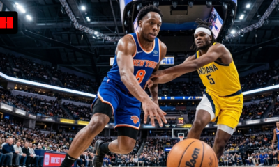 A low-angle, wide-lens action shot of New York Knicks forward OG Anunoby (number 8) dribbling a Wilson basketball past Indiana Pacers forward Jarace Walker (number 5) in a crowded NBA arena.