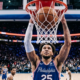 Philadelphia 76ers player finishes a powerful dunk during an NBA game at Xfinity Mobile Arena with the crowd reacting in the background.