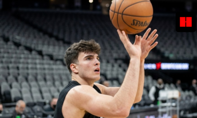 A young male basketball player in a black jersey focused on the hoop while releasing a jump shot in a large, empty arena.