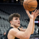 A young male basketball player in a black jersey focused on the hoop while releasing a jump shot in a large, empty arena.