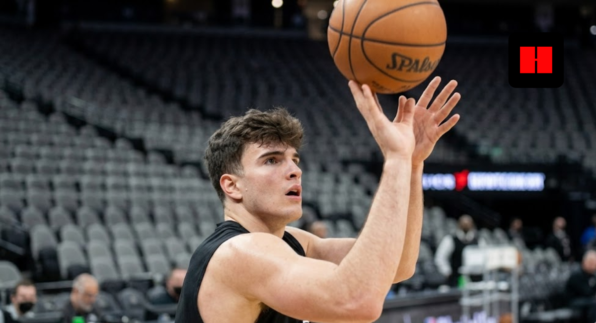 A young male basketball player in a black jersey focused on the hoop while releasing a jump shot in a large, empty arena.