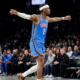 Shai Gilgeous-Alexander of the Oklahoma City Thunder celebrating on the court with arms outstretched, wearing a blue jersey and white headband during an NBA game.