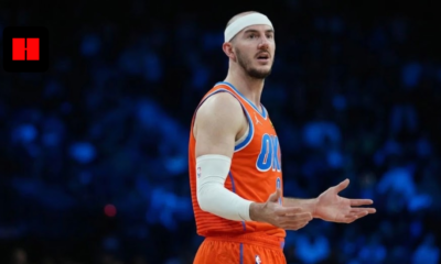 Alex Caruso of the Oklahoma City Thunder wearing an orange jersey and white headband, gesturing with his hands during a basketball game with a blurred arena background.