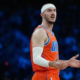 Alex Caruso of the Oklahoma City Thunder wearing an orange jersey and white headband, gesturing with his hands during a basketball game with a blurred arena background.