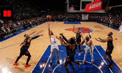 Wide-angle view of NBA basketball game showing Atlanta Hawks player attempting a rebound against New York Knicks defenders under the hoop in a packed arena