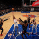 Wide-angle view of NBA basketball game showing Atlanta Hawks player attempting a rebound against New York Knicks defenders under the hoop in a packed arena
